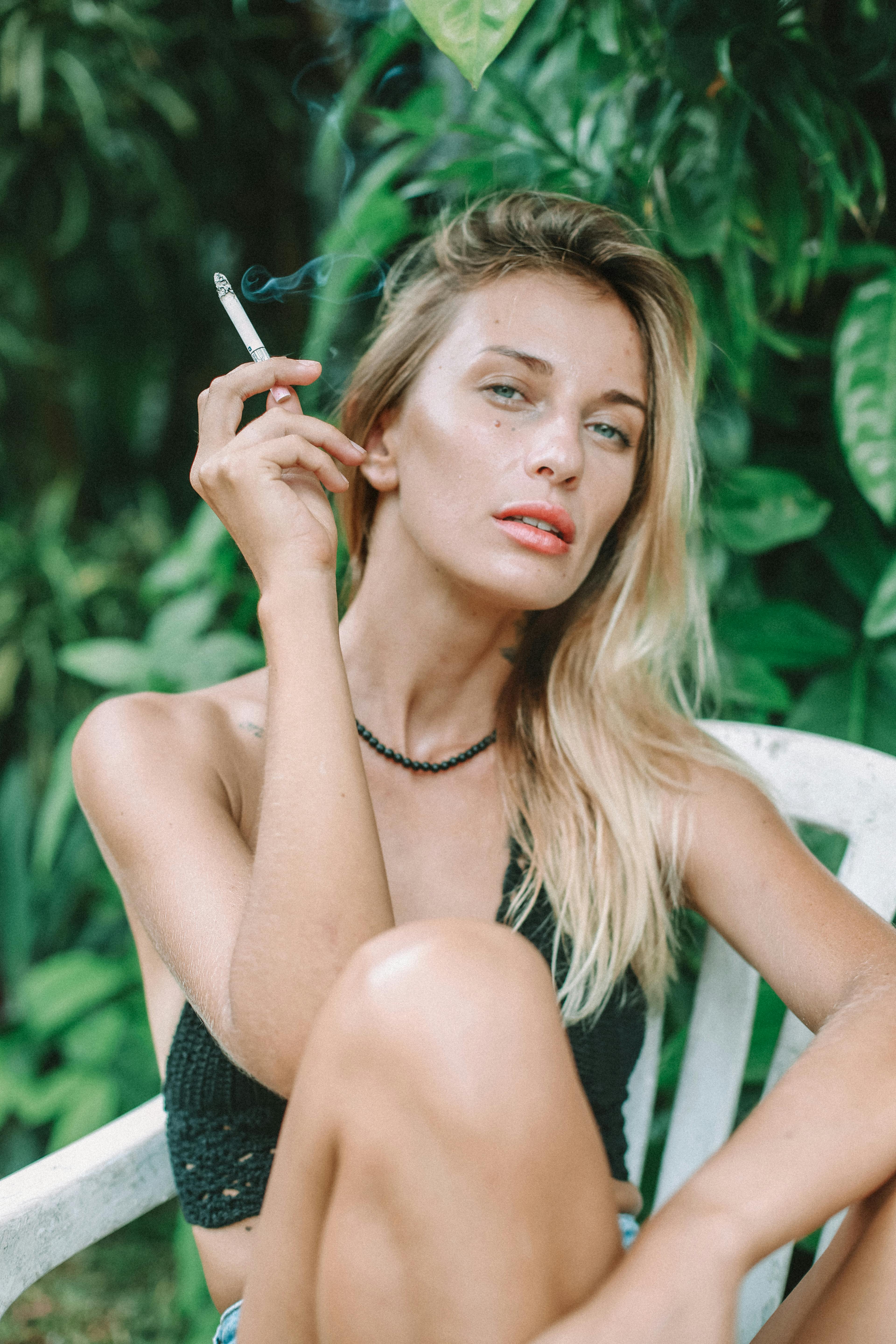 Blonde woman sits in garden holding cigarette, serene outdoor portrait.