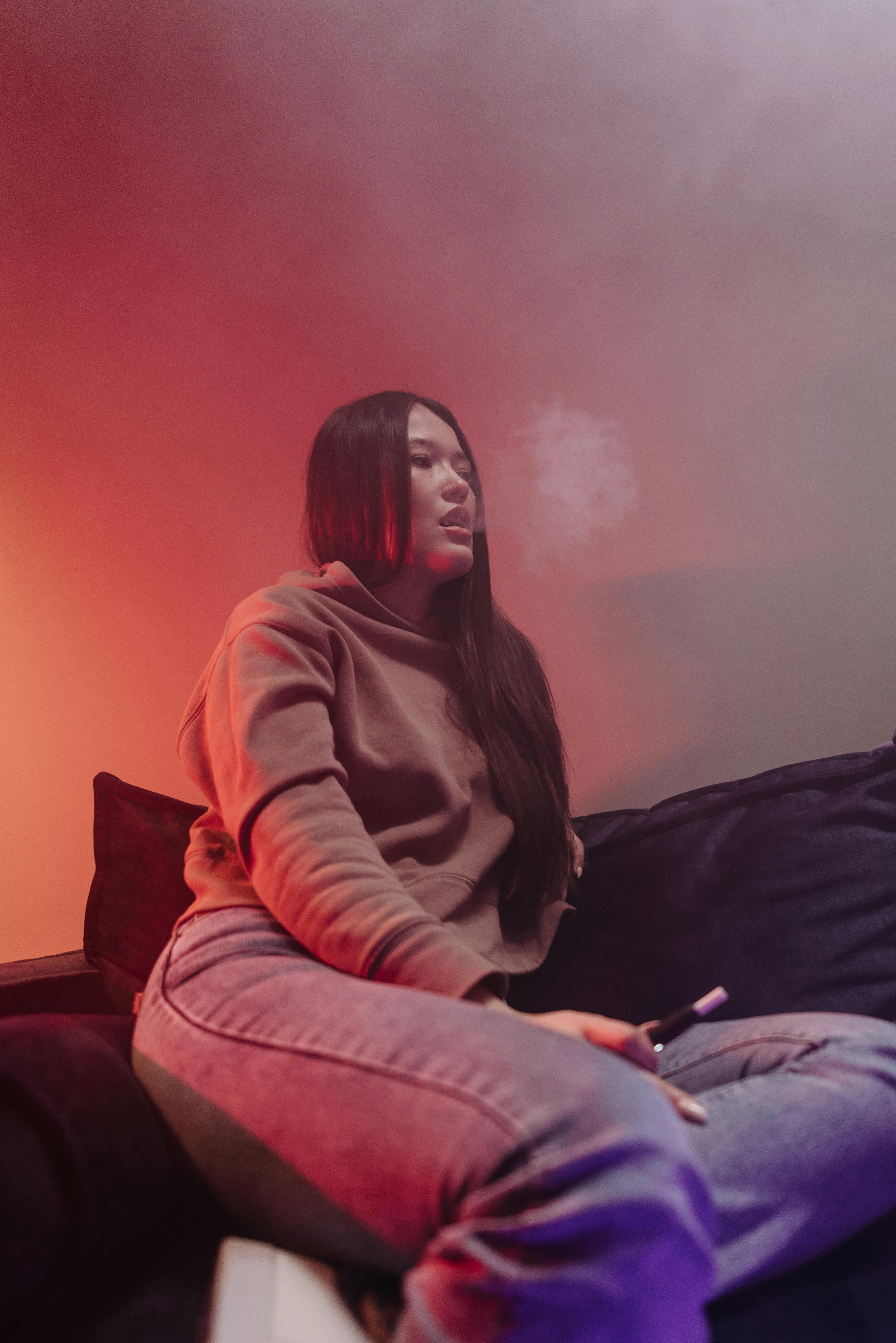Stylish young woman sitting indoors, exhaling vape smoke in a cozy setting with a warm ambiance.