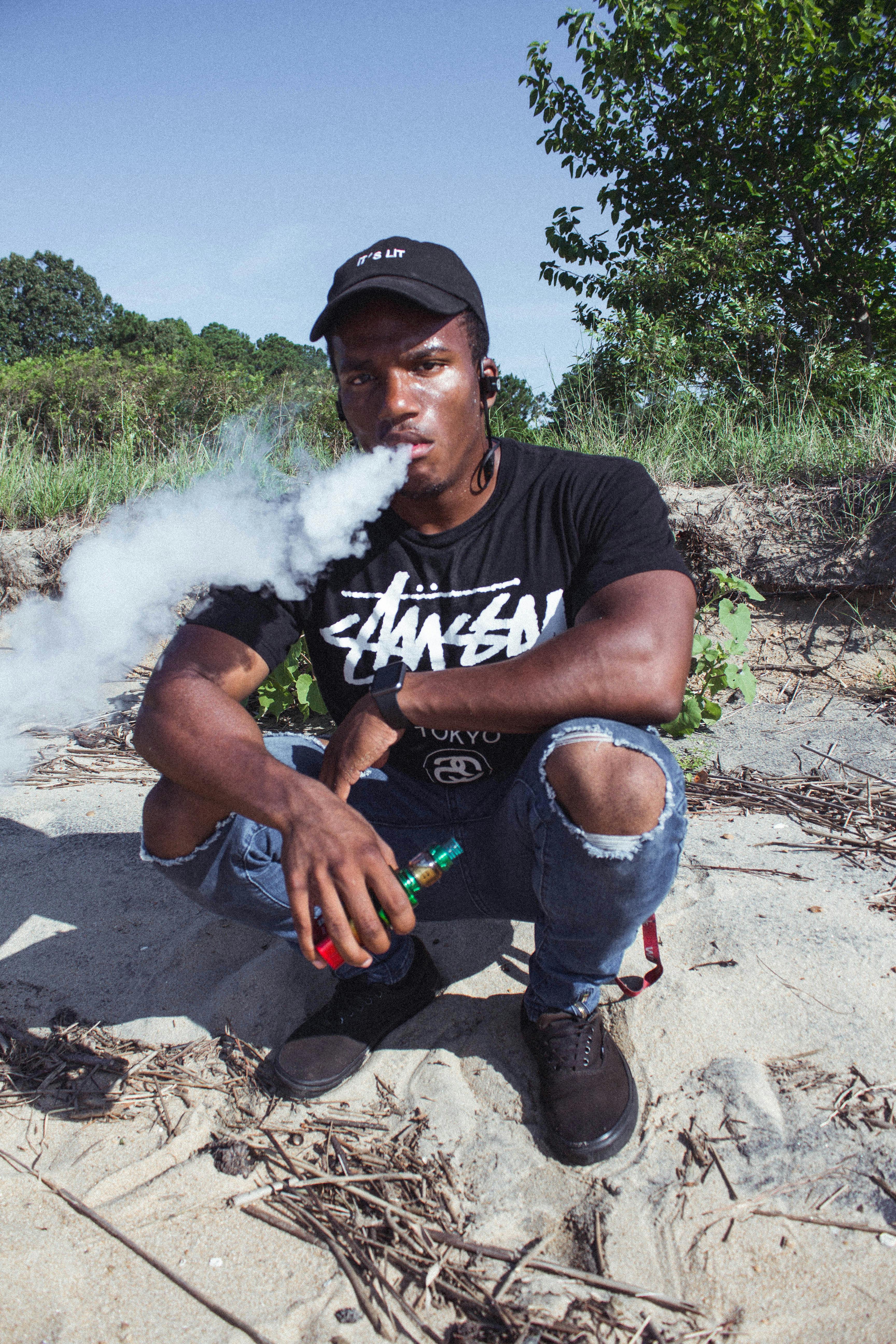 Young man in casual attire vaping outdoors near a natural landscape.